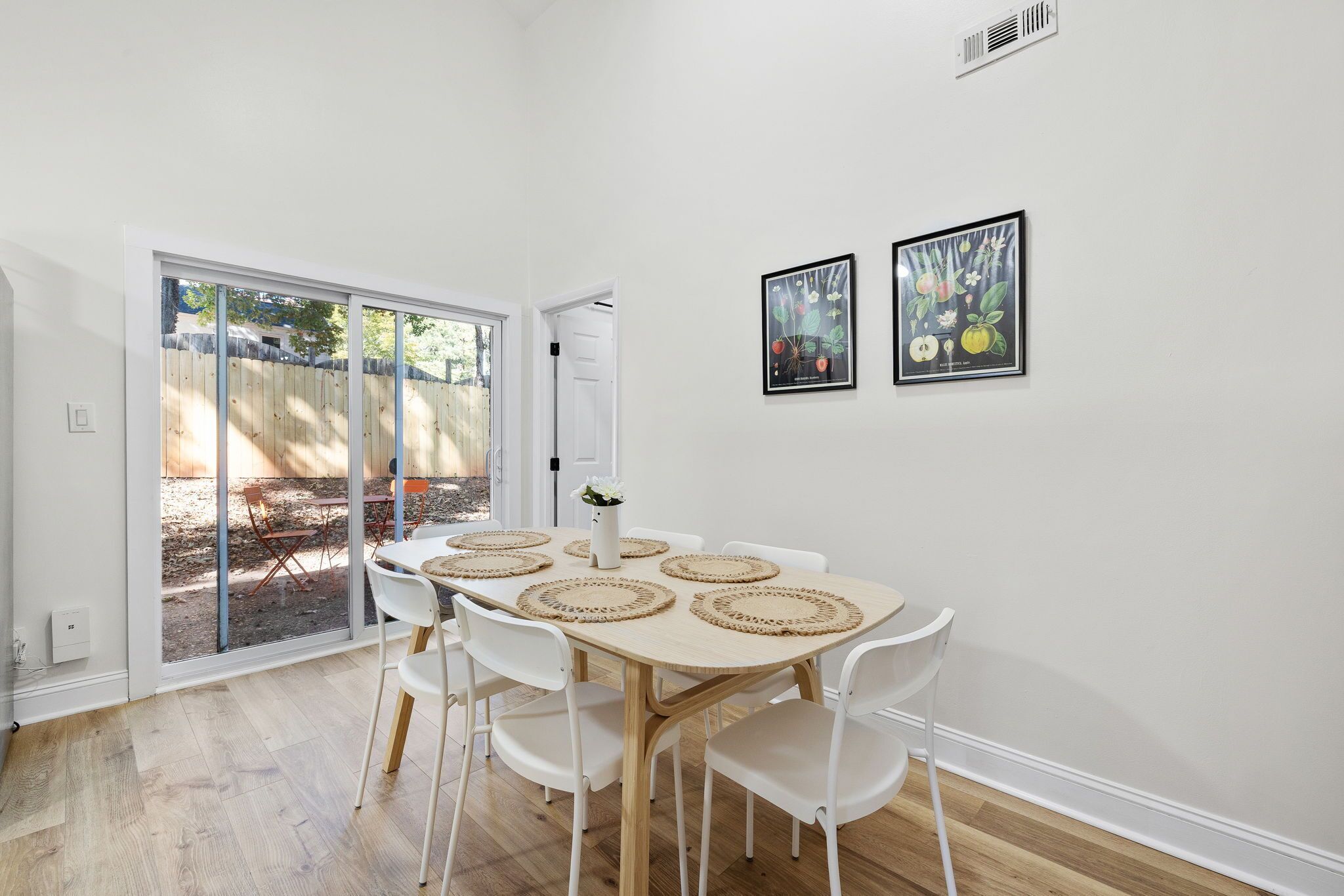 A dining area with a round table, white chairs, and woven placemats. There are two framed artworks on the wall and a sliding glass door leading to an outdoor area.