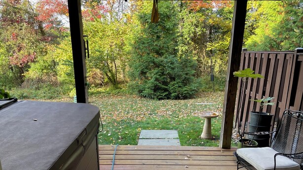 Eine Holzterrasse mit Whirlpool, einer überdachten Sitzecke und Blick auf einen Garten mit Bäumen und Vogelbad.