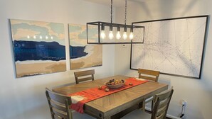 Ein Essbereich mit einem Holztisch, vier Stühlen, einer roten Tischmatte und einer Schale mit Snacks. An der Wand befinden sich zwei abstrakte Gemälde und eine Karte.