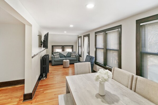 A modern living room with a sofa, a dining table with a vase of flowers, and a kitchen area in the background.