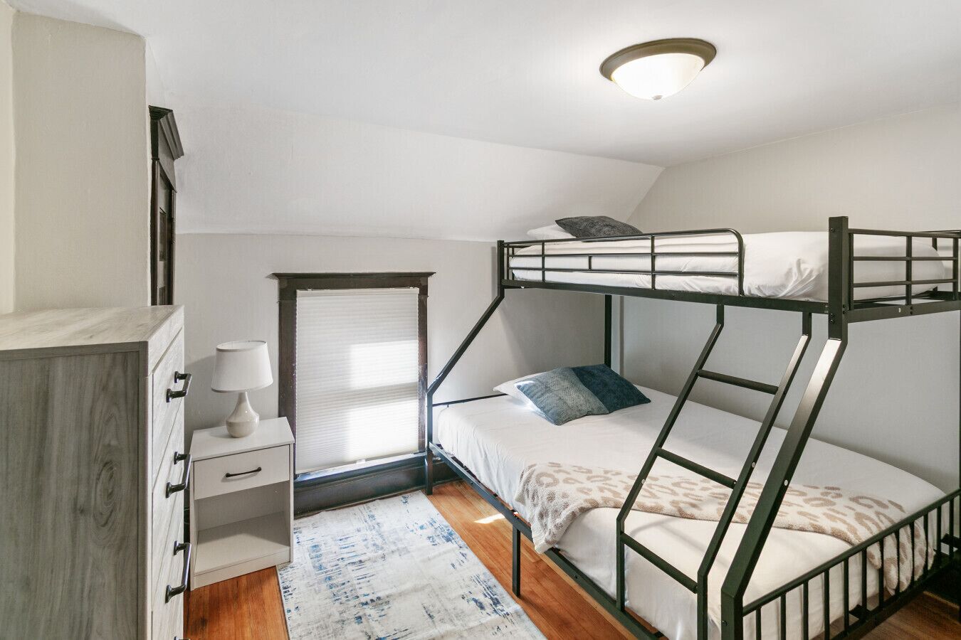 Una habitación tipo dormitorio con literas, un escritorio con cajones, una lámpara y una ventana con persianas.
