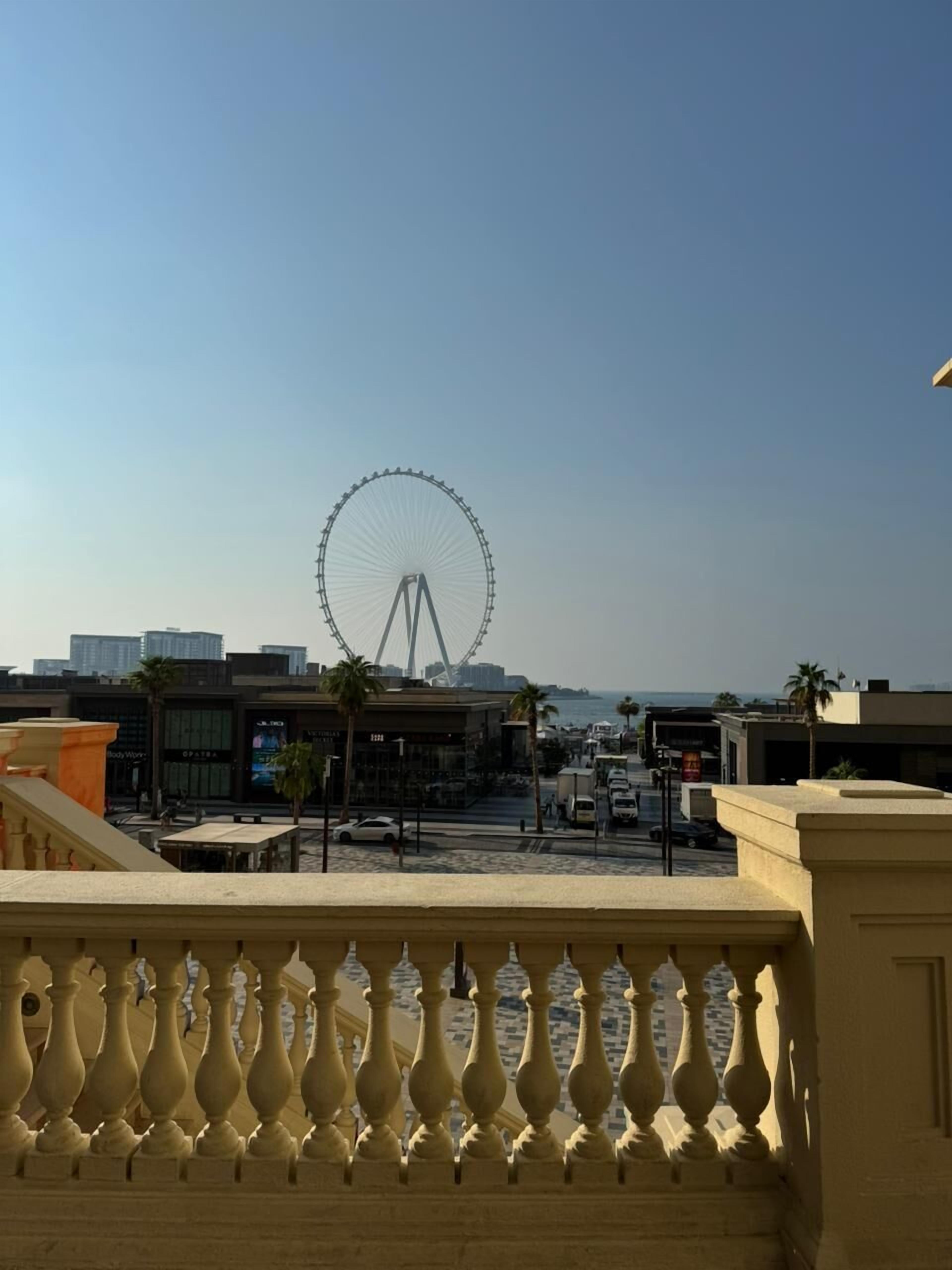 Une vue sur un paysage urbain, avec une grande roue bien visible au fond, des bâtiments modernes et un balcon doté de balustrades.