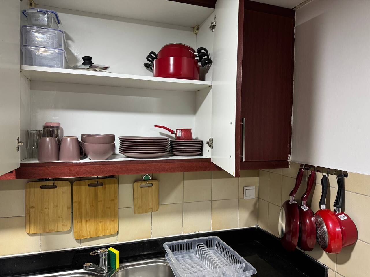 A kitchen with white cabinets, a wooden countertop, and a black sink. There are red pots hanging on the wall, a wooden cutting board, and a plastic dish rack.