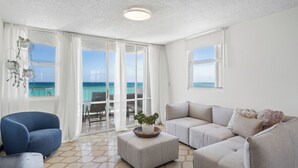 A spacious living room with a large window offering a view of the sea, a blue armchair, a grey sofa, and a white decorative bowl on a matching ottoman. - Oceanfront Terrace Communal Pool AC (Carolina)