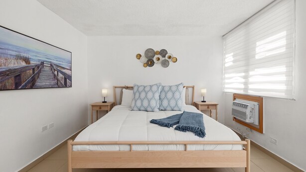 Une chambre à coucher avec un lit en bois, du linge de lit blanc, un tableau sur le thème de la plage, des éléments de décoration murale et une fenêtre avec des stores.