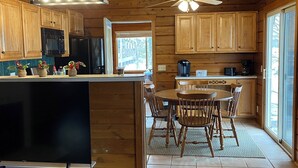 A wooden cabin kitchen with a dining area, wooden cabinets, a ceiling fan, and a large window. - Rustic UP Waterfront Cabin on Huron Bay (L'Anse)