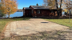 A rustic cabin with a gravel driveway leading to a wooden door, surrounded by autumn foliage and a lake view. - Rustic UP Waterfront Cabin on Huron Bay (L'Anse)