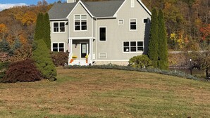 A two-story house with a front porch, a chimney, and a view of a forested hillside. - Luxurious Lakefront Retreat –  Sleeps 11 (Newtown)