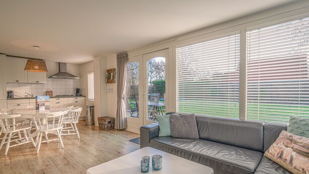 A modern kitchen with white cabinets, a dining table with chairs, and a grey sofa with cushions. - Cozy Detached Bungalow for 4 Persons (De Koog)