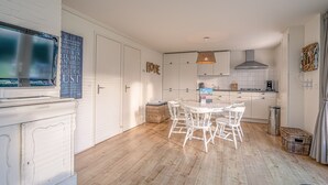A modern kitchen with white cabinets, a dining table with chairs, and a television mounted on the wall. - Cozy Detached Bungalow for 4 Persons (De Koog)