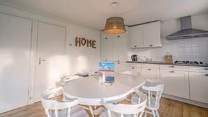 A modern kitchen with white cabinets, a round dining table with white chairs, a wicker pendant light, and a 'HOME' sign on the wall. - Cozy Detached Bungalow for 4 Persons (De Koog)