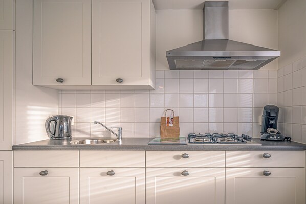 A kitchen with white cabinets, a stainless steel sink, a kettle, a paper bag, and a coffee maker. - Cozy Detached Bungalow for 4 Persons (De Koog)