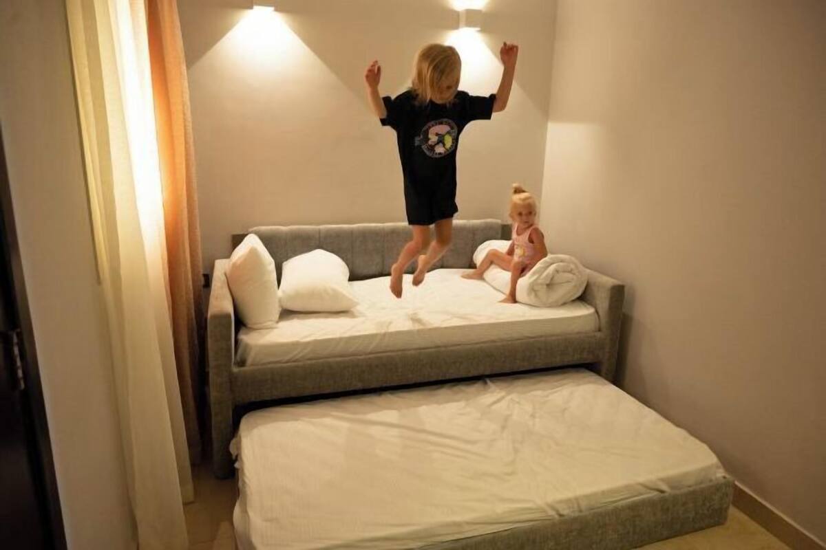 A child jumping on a bed in a room with a sofa and a wall-mounted light fixture.