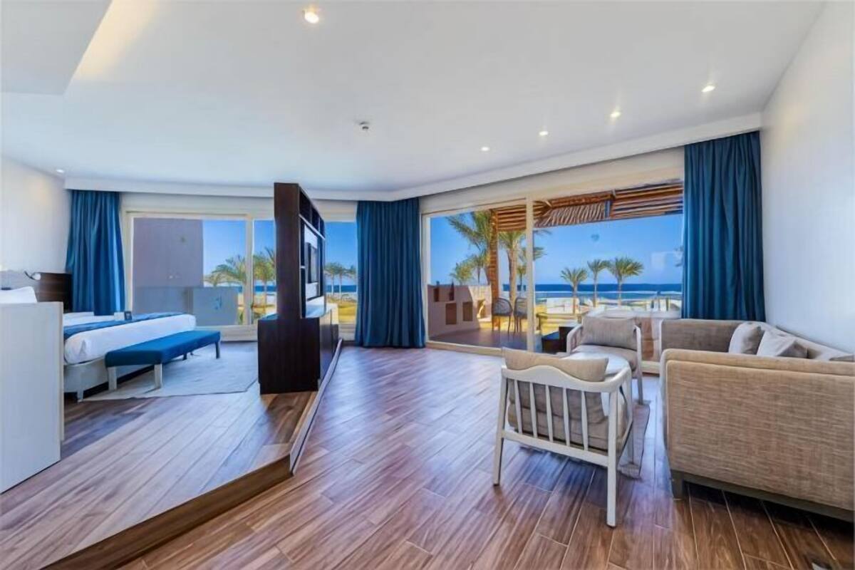 A hotel room with a bed, a sofa, a TV, a dining table, and a view of the beach.