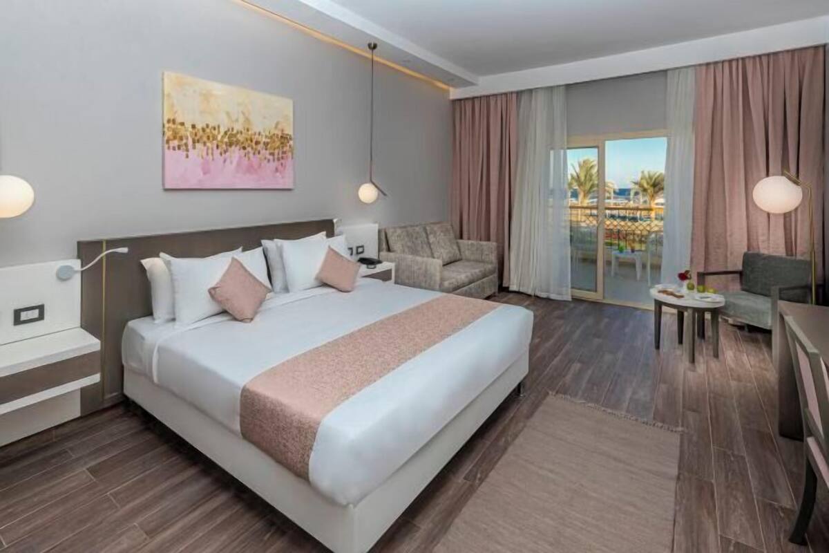 A modern hotel room with a large bed, a sofa, a small table, and a view of the outdoors.