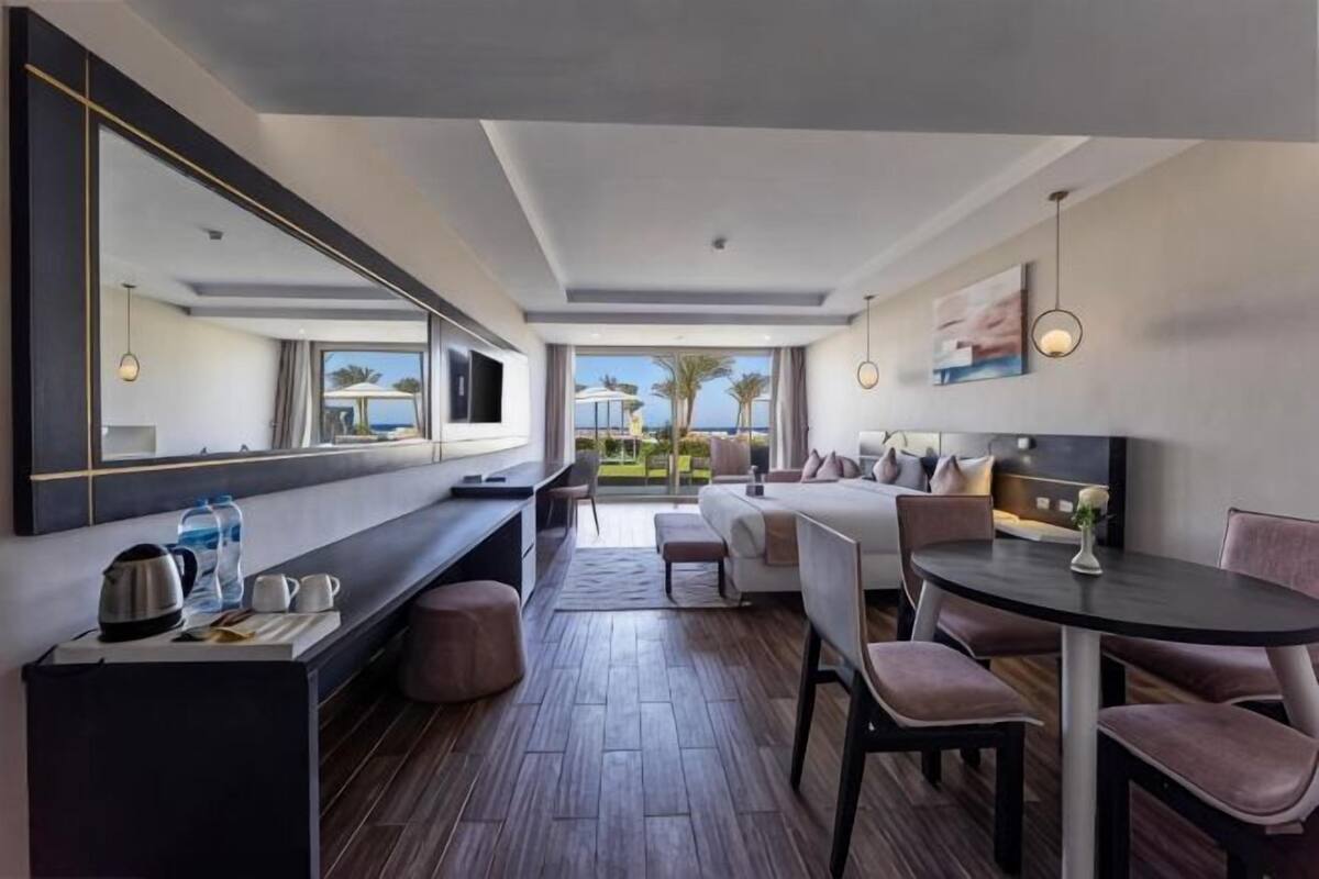 A modern hotel room with a large bed, a desk, a dining area, and a view of palm trees.