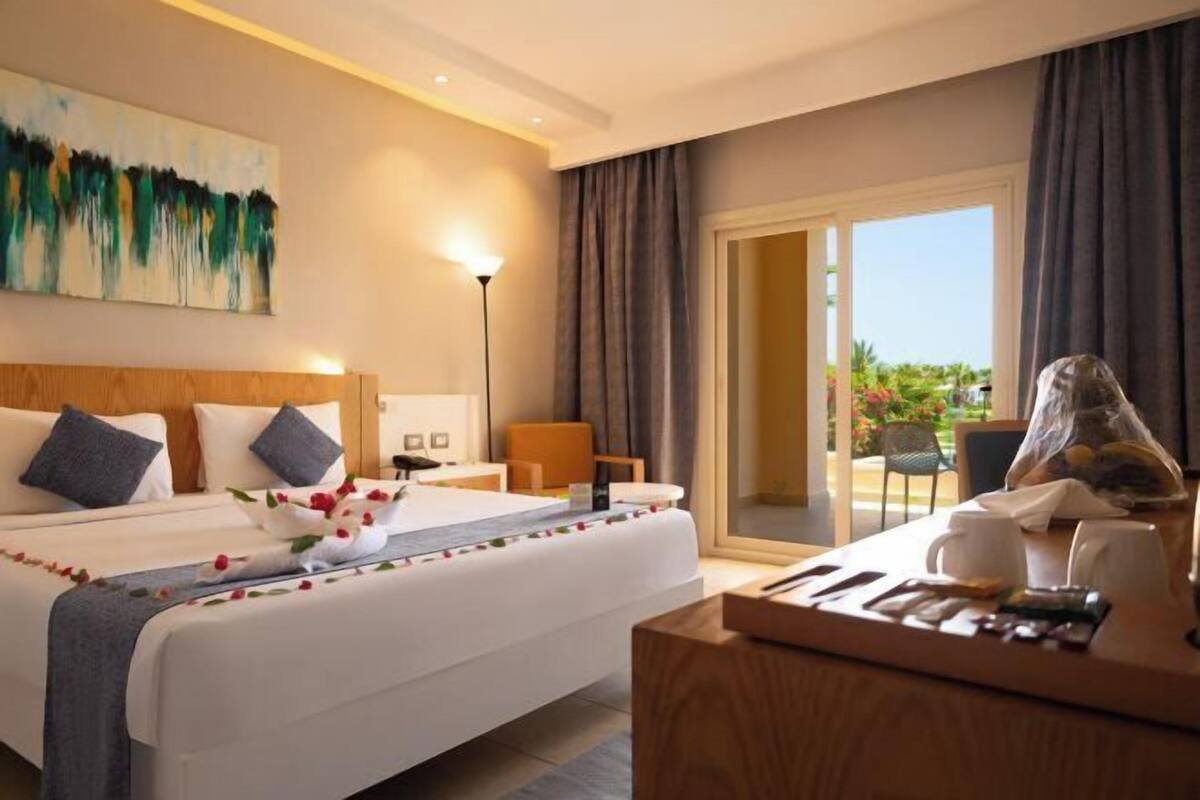 A hotel room with a bed, a desk with a lamp, a chair, and a view of the outdoors.