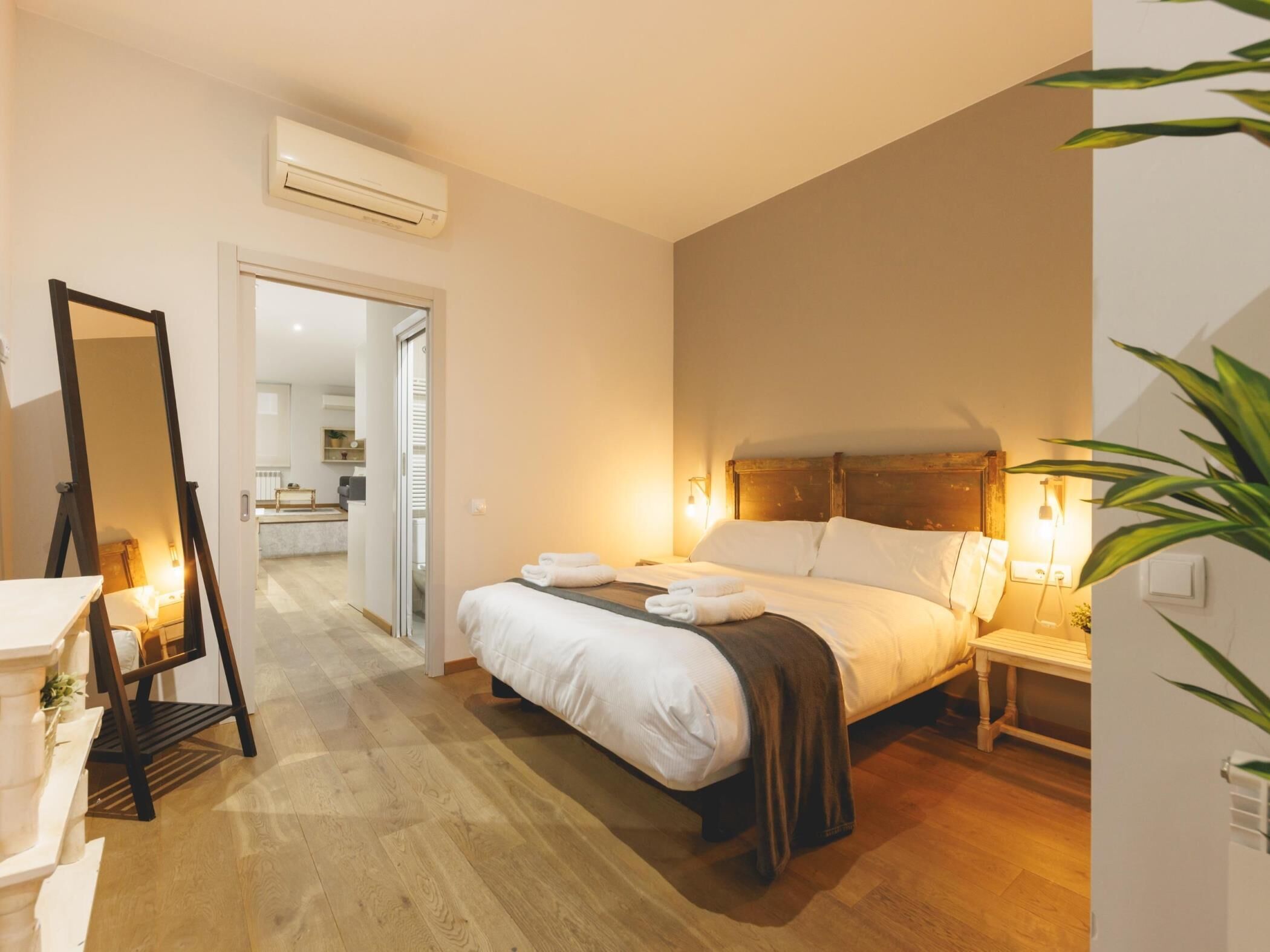 Een slaapkamer met een bed, nachtkastje en een spiegel. Er is een deur naar een badkamer en een gang.