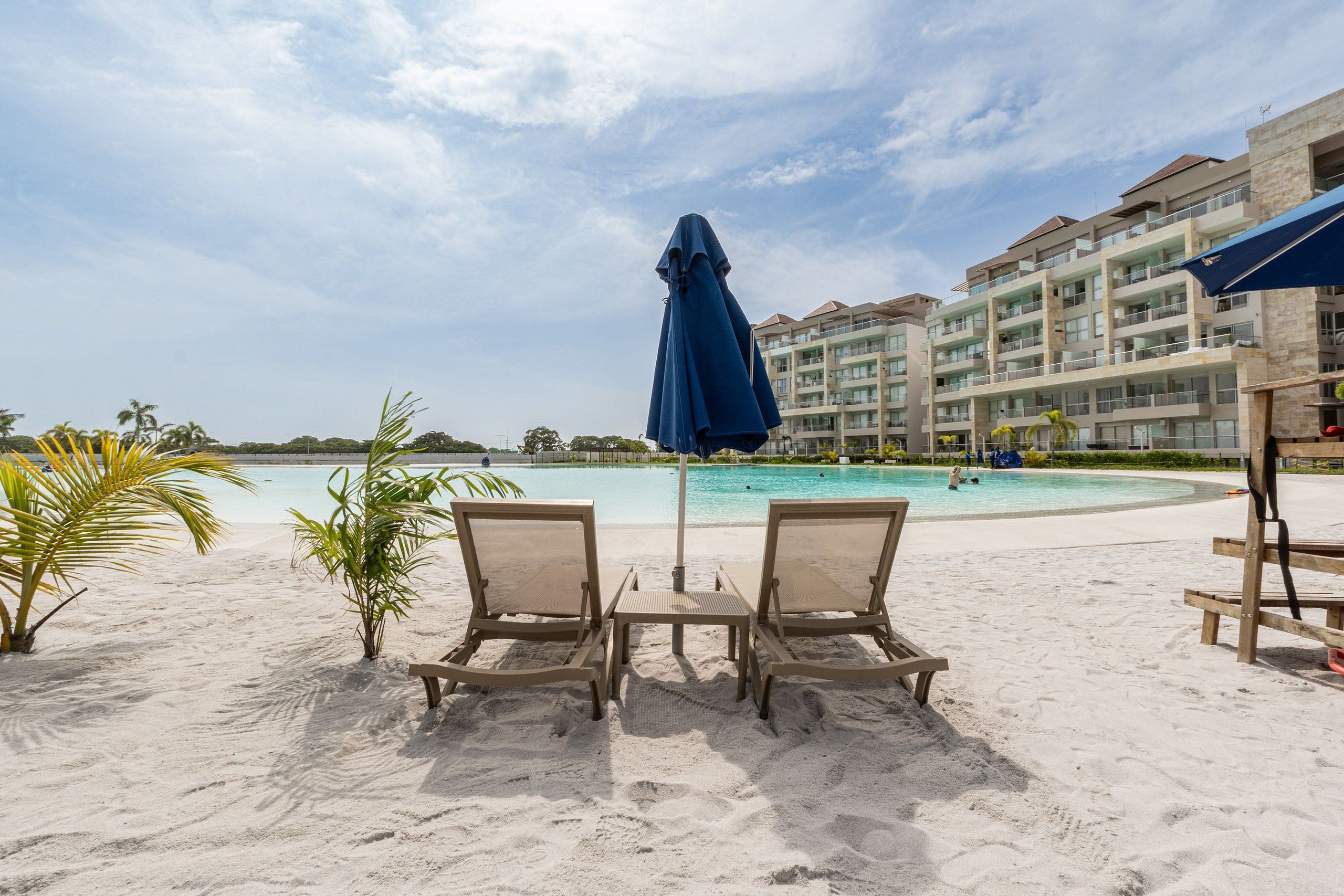 Una playa con dos sillas de descanso y una mesa, piscina y un edificio con balcones al fondo.
