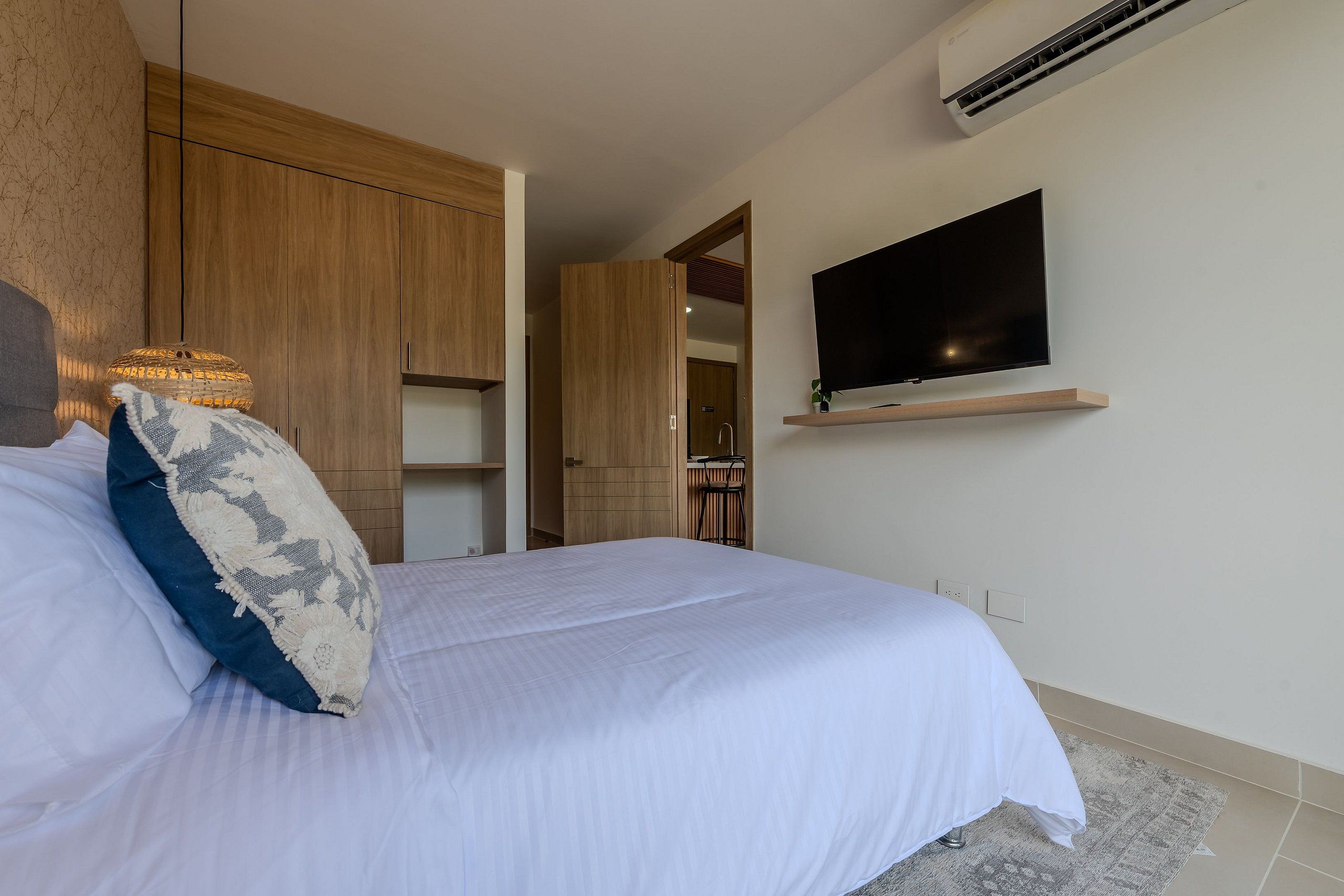 Una cama bien tendida con una colcha blanca y un cojín decorativo, un televisor montado en la pared y muebles de madera al fondo.