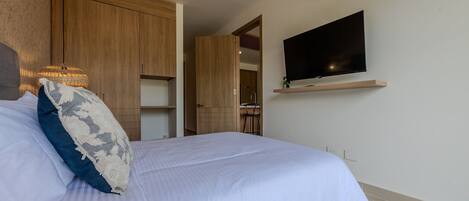 Una cama bien tendida con una colcha blanca y un cojín decorativo, un televisor montado en la pared y muebles de madera al fondo.
