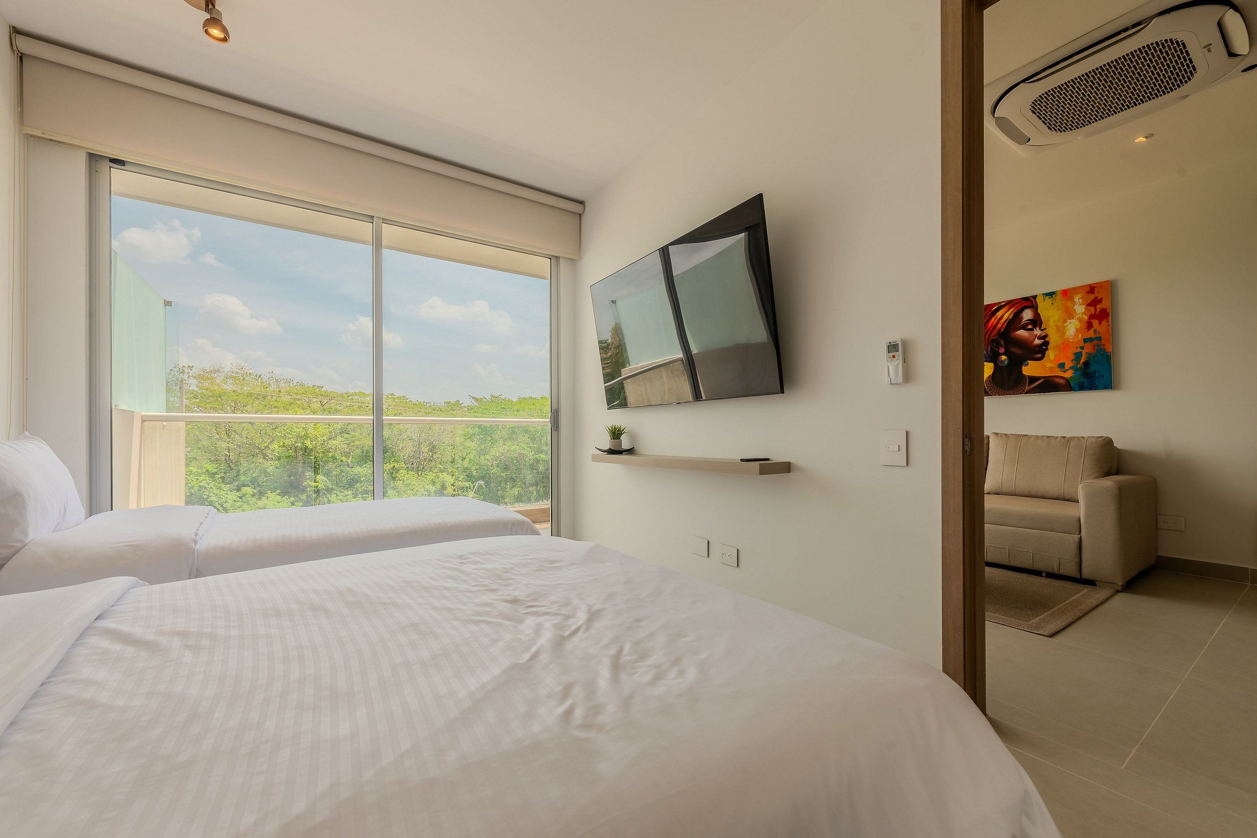 Un dormitorio con dos camas, un televisor montado en la pared, un sofá y un amplio ventanal con vistas al follaje.