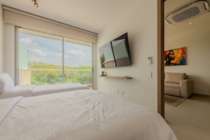 Un dormitori amb dos llits, un televisor fixat a la paret, un sofà i una finestra gran amb vista a vegetació.
