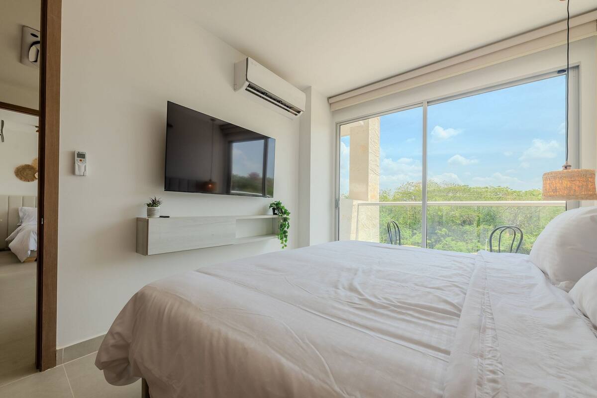 Quarto com cama grande, TV instalada, ar condicionado e janela com vista para vegetação.