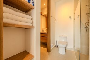 A modern bathroom with a glass shower enclosure, a toilet, and a wooden shelf with towels. - Apartamento premium en Baia Kristal (Cartagena)