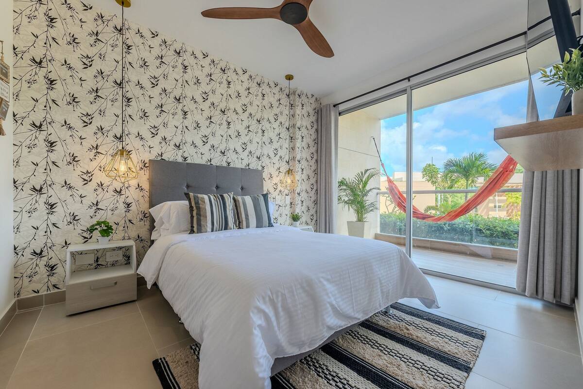 Um quarto com cama, ventilador de teto, mesa de cabeceira, janela com vista para uma rede vermelha lá fora e papel de parede estampado.