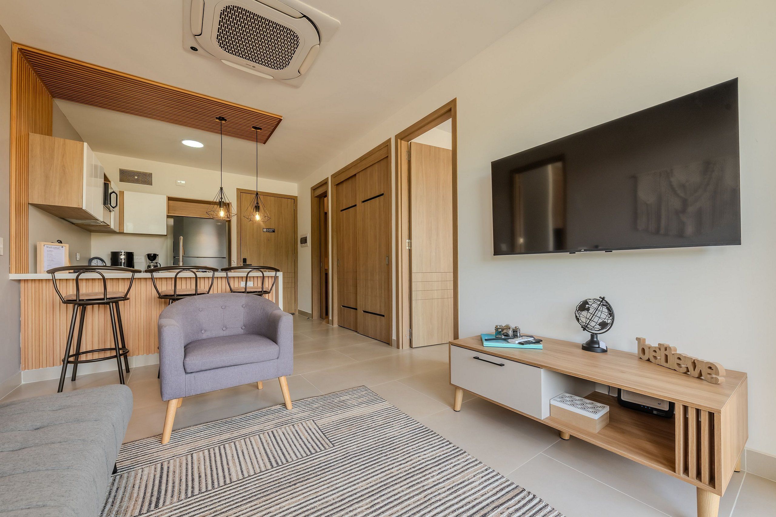 A modern living room with a grey sofa, wooden TV stand, and a kitchen area with wooden cabinets and a bar stool.