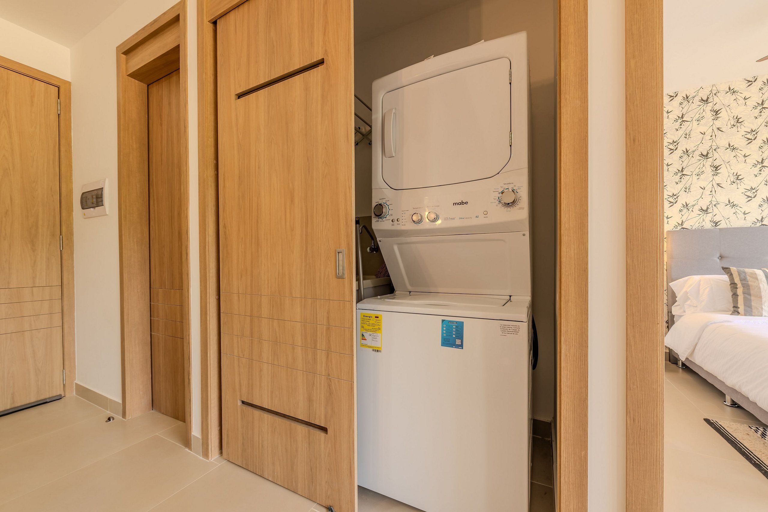 Un lavadero con lavadora de carga frontal, puertas de madera, y en la habitación contigua, una cama con ropa blanca.