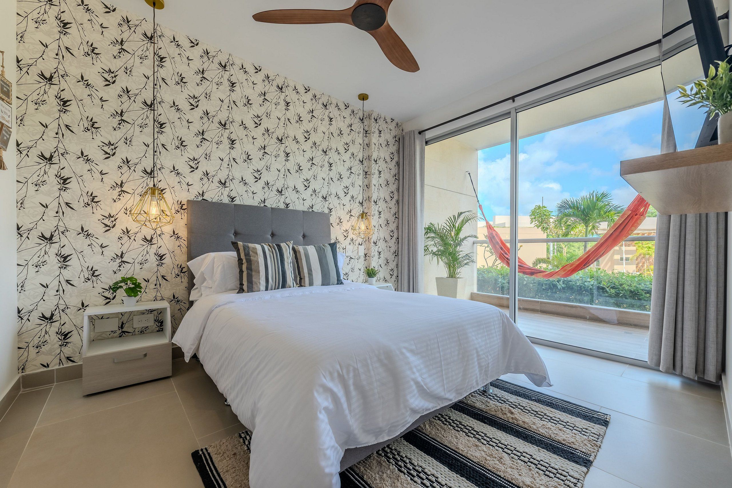 A bedroom with a bed, a ceiling fan, a nightstand, a window with a view of a red hammock outside, and a patterned wallpaper.