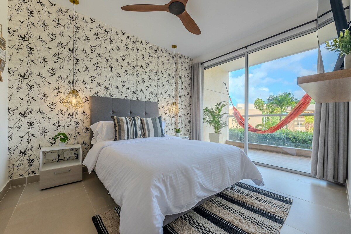 A bedroom with a bed, a ceiling fan, a nightstand, a window with a view of a red hammock outside, and a patterned wallpaper.