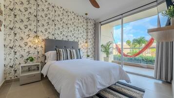 A bedroom with a bed, a ceiling fan, a nightstand, a window with a view of a red hammock outside, and a patterned wallpaper.