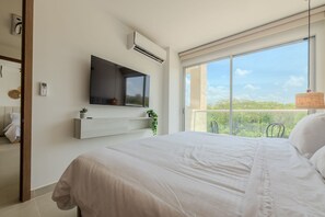 A bedroom with a large bed, a mounted TV, an air conditioner, and a window with a view of greenery. - Apartamento premium en Baia Kristal (Cartagena)