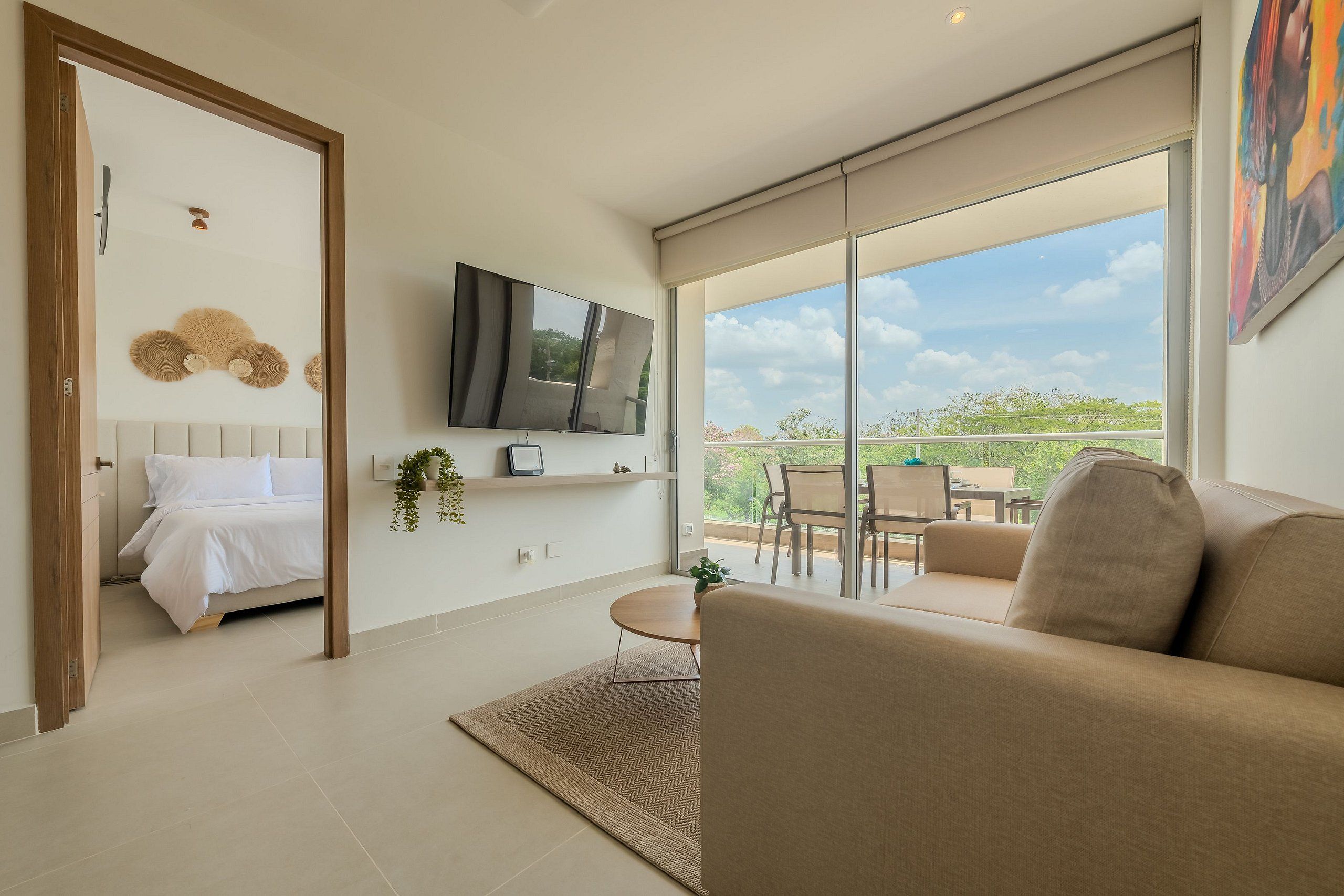 A modern living room with a sofa, a TV mounted on the wall, and a balcony with a view of greenery.
