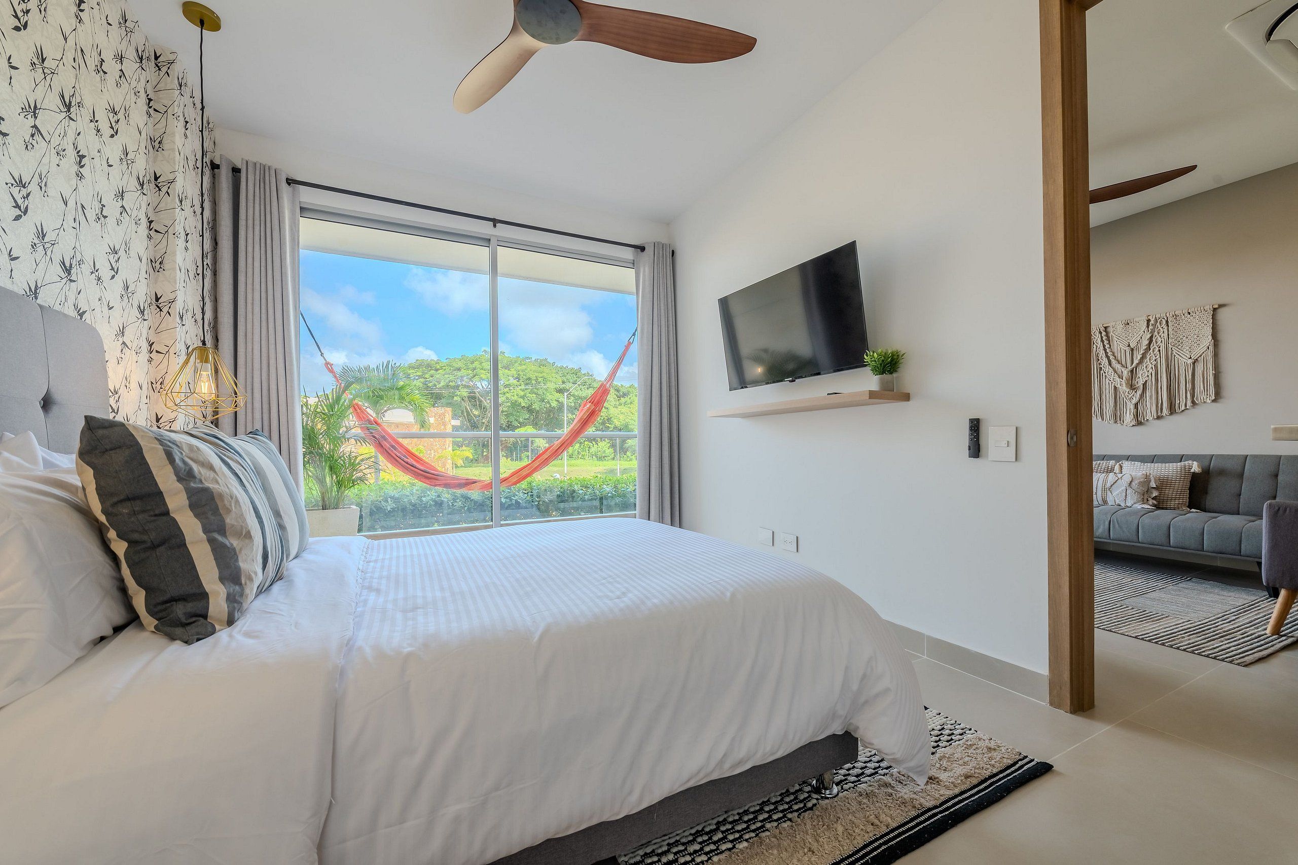 A modern bedroom with a large bed, a TV mounted on the wall, and a view of greenery outside.