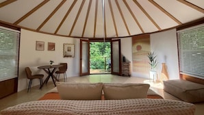 Une pièce avec un plafond haut, un lit au premier plan, un coin repas avec des chaises et une table, et une vue sur l’extérieur par une porte ouverte.