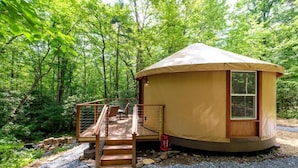 Une yourte circulaire, de plain-pied, dotée d’une terrasse en bois et d’une petite fenêtre, entourée d’une zone boisée.