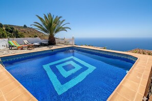 Una piscina con diseño geométrico, sillas de descanso, una palmera y vista al mar.