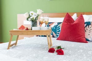 Een bed met een wit gebedrukte sprei, een houten dienblad met een stuk watermeloen, twee glazen van een helder drankje en een vaas met witte bloemen.