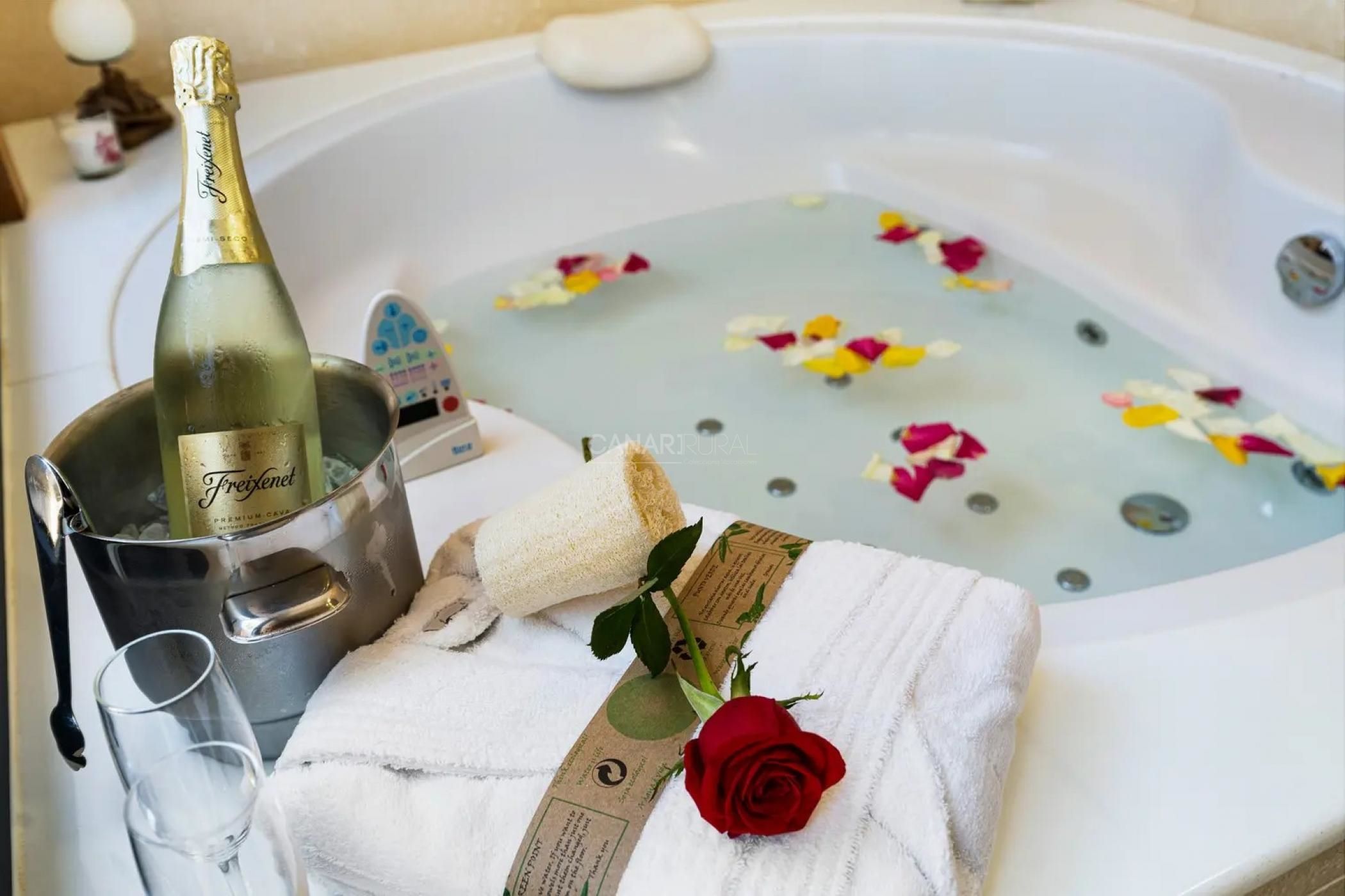A bathtub with rose petals, a bottle of champagne, a towel, and a glass.