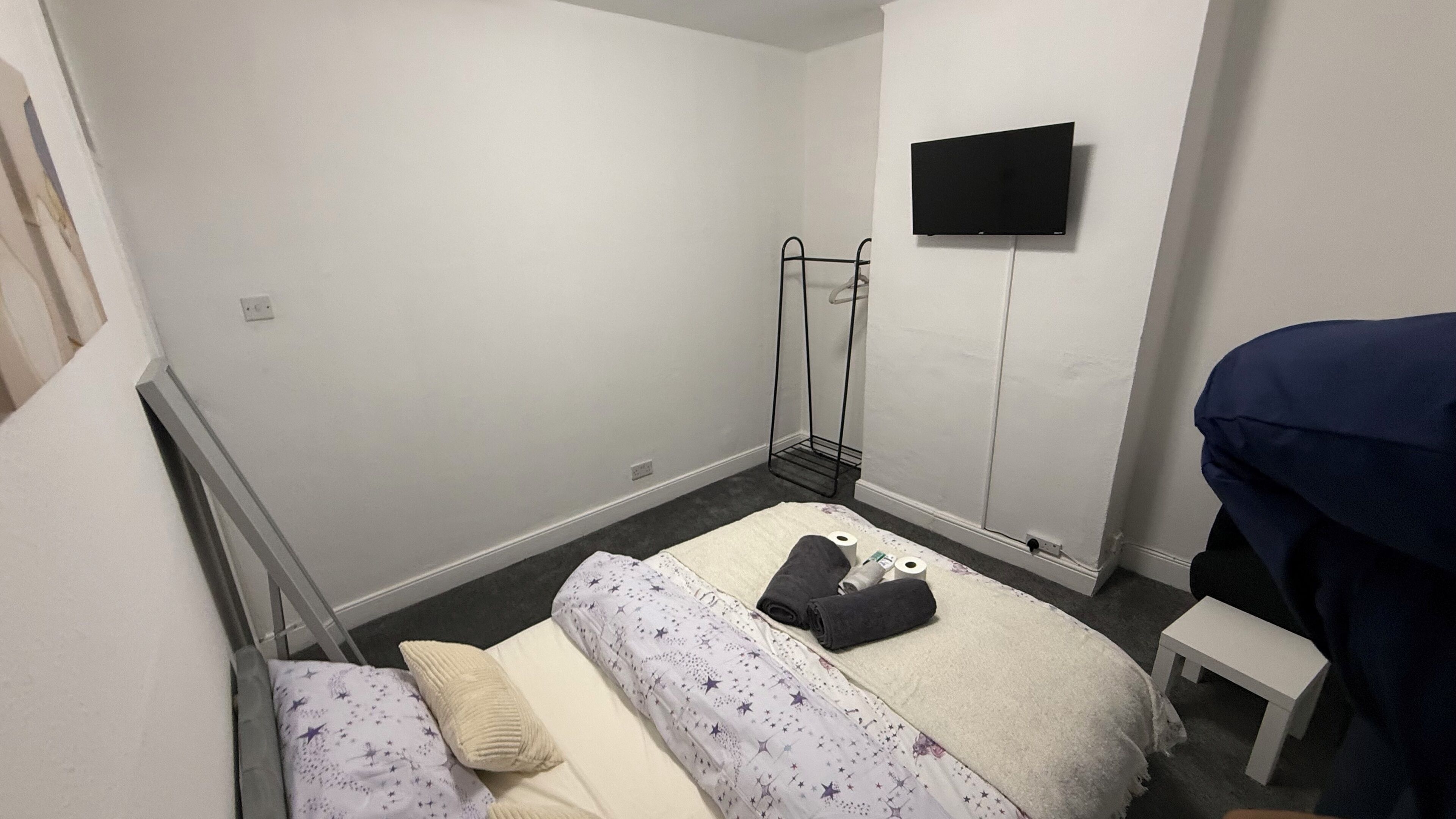 A small, sparsely furnished room with a single bed, a small table, a TV mounted on the wall, and a radiator.