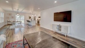 A spacious living room with a large TV, wooden flooring, and a modern kitchen in the background.