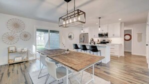 A modern kitchen with a wooden dining table, white chairs, and a hanging light fixture.