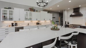 Uma cozinha moderna com armários brancos, uma ilha central com banquetas, um lustre e um fogão.