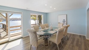 A dining area with a glass table and six chairs, large windows with a view of a body of water, and a wooden deck outside. - 2 Lakefront Homes: Wide Decks, Great View (Gravois Mills)