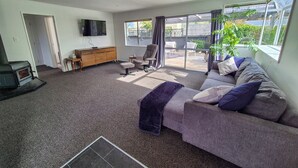 A living room with a sofa, armchair, and a television. There is a fireplace, a dining table, and a window with curtains. - Comfy family home, stay at our retreat on Brittan St! (Hokitika)