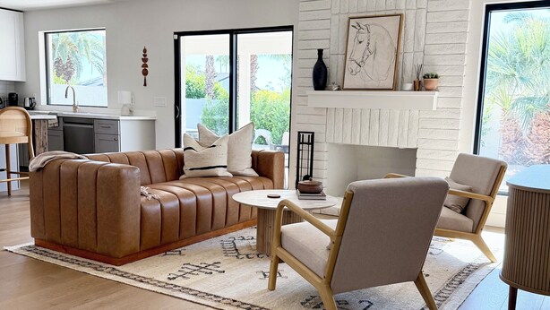 A modern living room with a leather sofa, a round coffee table, and a beige armchair. There is a fireplace with a framed artwork above it, and a kitchen area is visible in the background. - Peaceful 3BD/2BA Dog-Friendly Paradise Posada (Scottsdale)